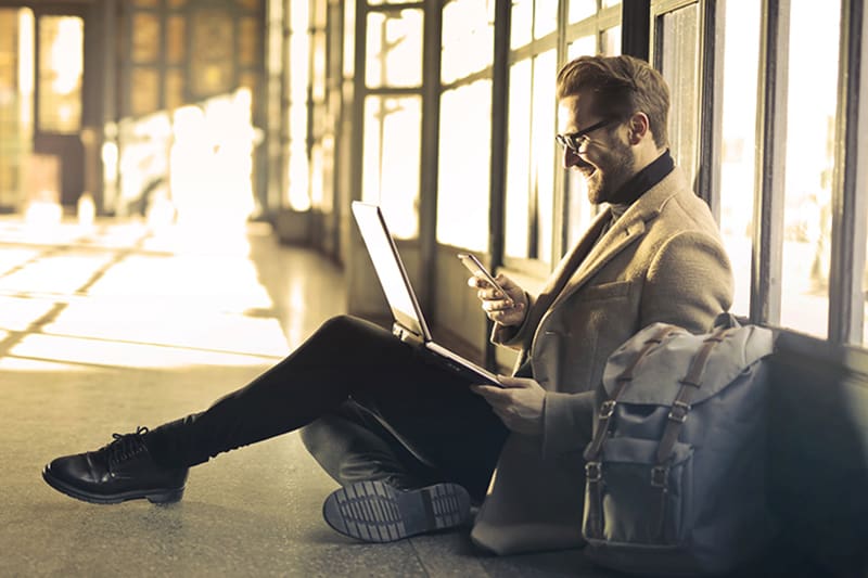 man sitting near window holding phone and laptop