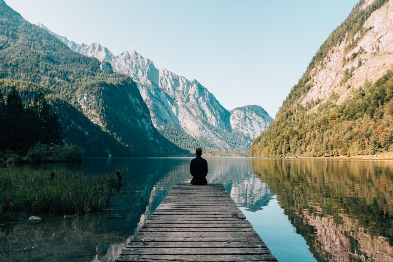 man in black jacket sitting on wooden dock looking at mountain