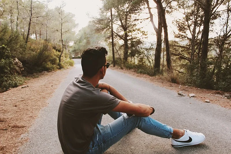 man sitting on gray concrete road surrounded with trees