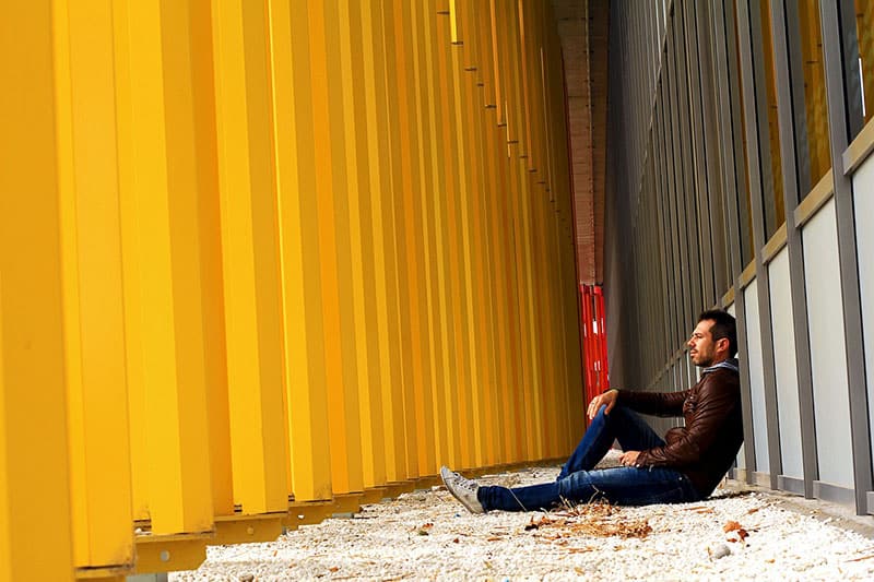 man sitting on walkway in between yellow columns and gray steel building