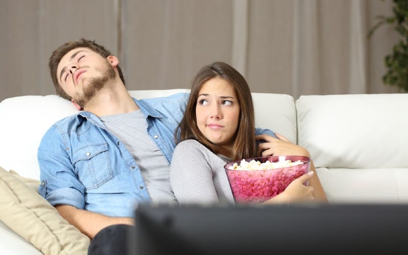 Man sleeping on couch near bored woman watching tv holding a popcorn bowl