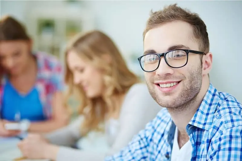 man smiling wearing eyeglasses with two women in a blurred distant from him