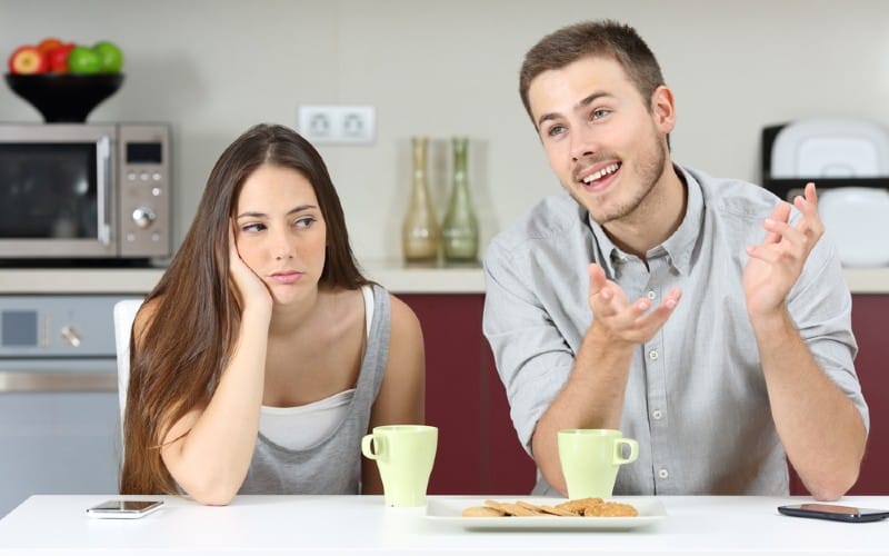 Hombre hablando con una mujer aburrida sentado a su lado delante de una mesa con tazas de café y galletas.