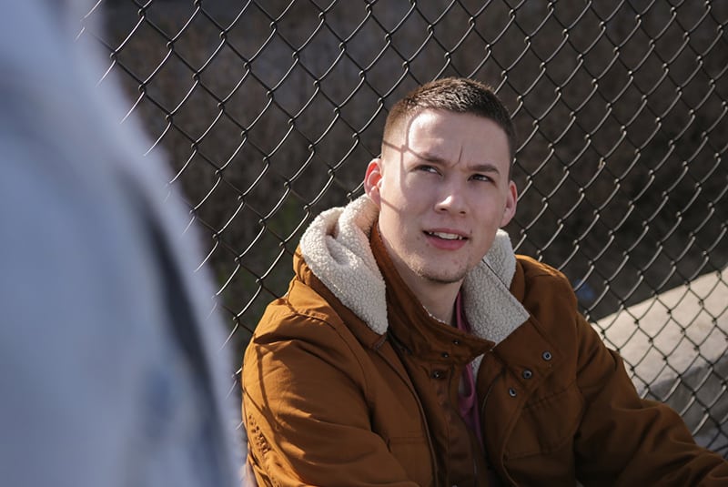 man wearing brown jacket sitting near gray link fence