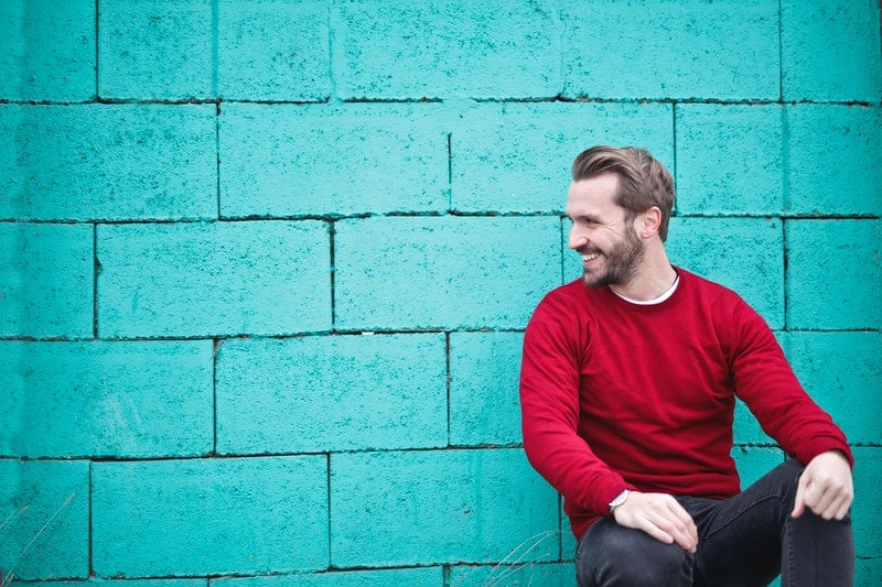 man wearing red sweatshirt and black pants leaning on the teal brick wall
