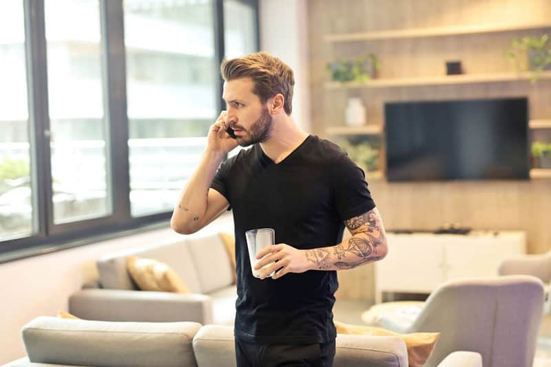 man whispers on phone while holding glass inside a house 