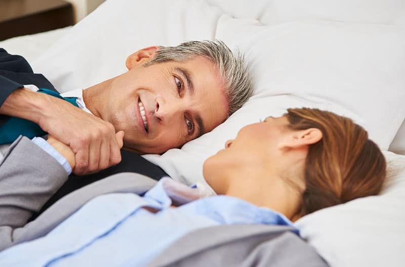 older man and woman lie down on bed on their office suit