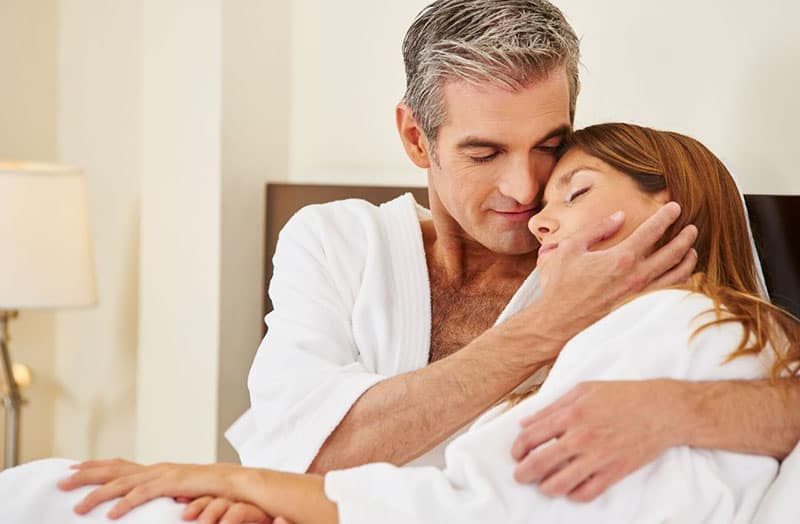 older man hugging young woman both in white robe in bed