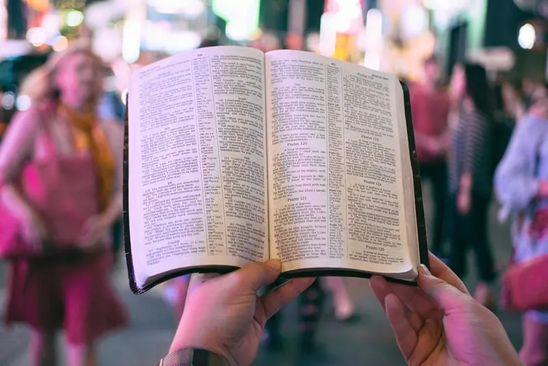 person holding bible on road with people walking on sidewalk