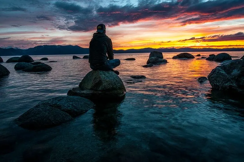 person sitting on rock on water facing a sun setting