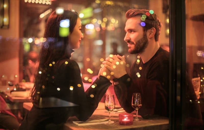 photo of a couple holding hands in a dinner set up with lights reflecting on the glass window beside them