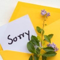 I am sorry message card handwriting in yellow envelope with purple flower arrangement on background white wooden