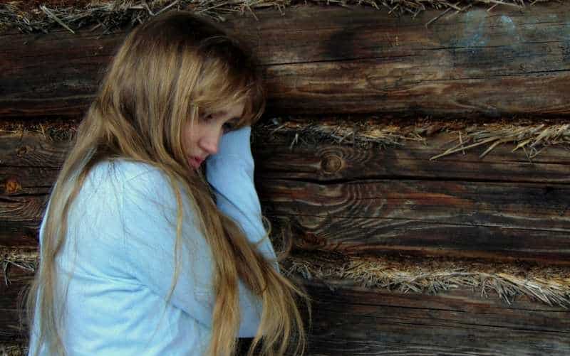 Sad blonde woman with long hair wearing blue top standing near wooden wall