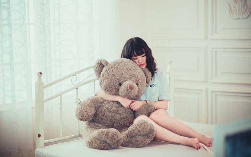 Sad woman holding big teddy bear while sitting on the bed