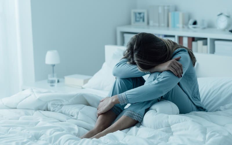 Sad woman wearing pajama sitting in bed with her head on her knees