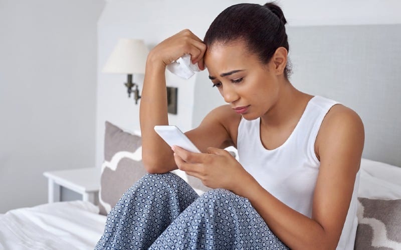 Triste mujer joven sentada en la cama y los mensajes de texto en el teléfono con tisue en una mano durante el día