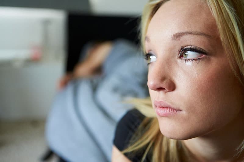 sad young woman in focus with a person sleeping on bed at a distance in blur image