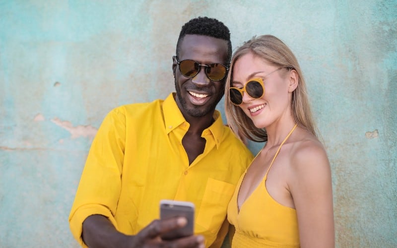 Smiling man in yellow shirt standing beside smiling woman looking at a phone