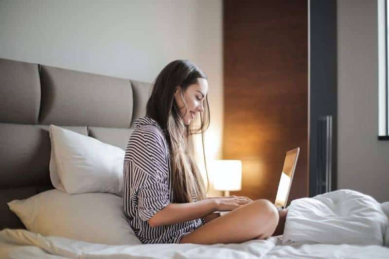 donna sorridente con un top nero e a righe seduta su un letto mentre utilizza un computer portatile
