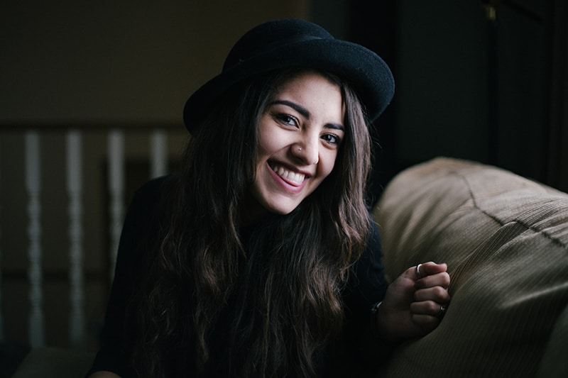 mulher sorridente com cabelo preto e chapéu preto sentada no sofá