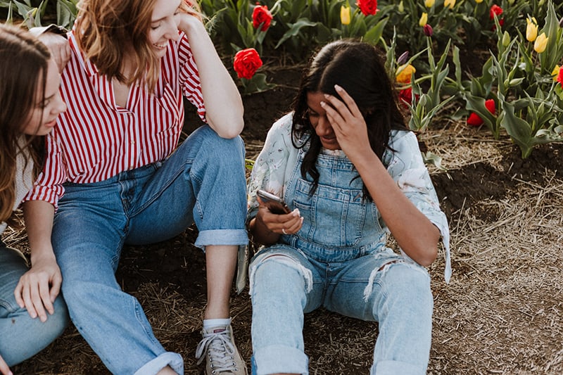 tres mujeres riendo mientras miran el smartphone cerca del campo de flores