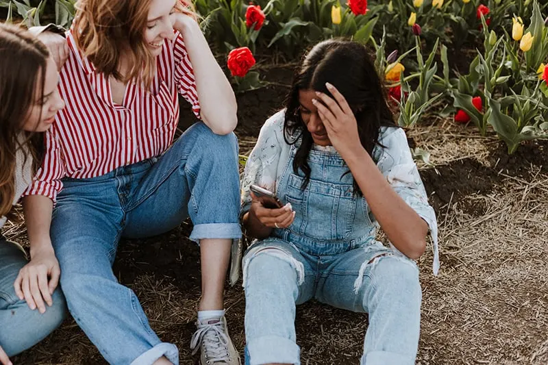 three women laughing while looking at smartphone near the flower field