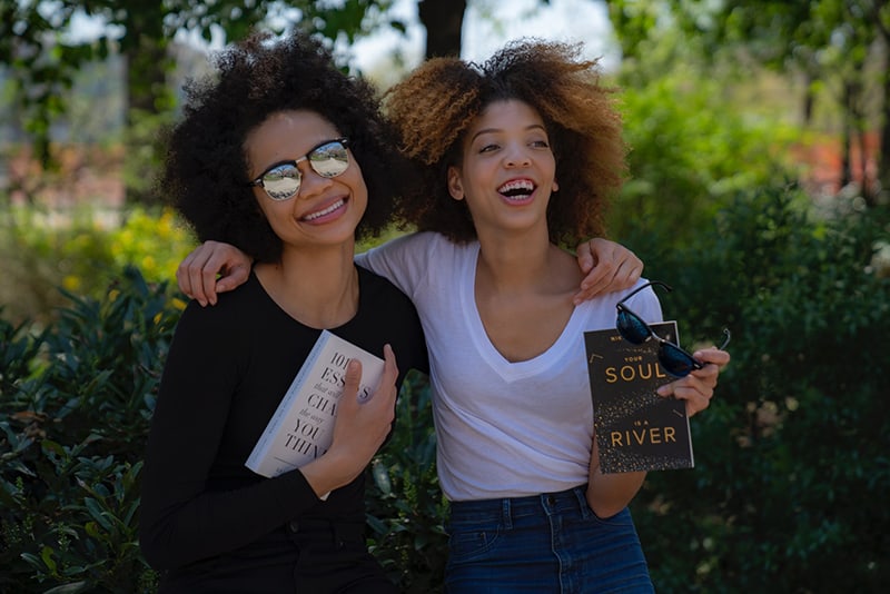 two smiling women while holding novel books surrounded with trees