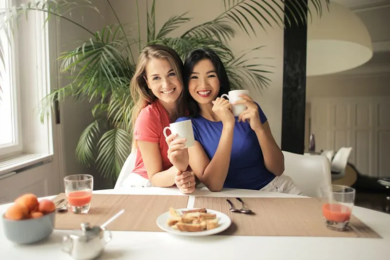 two women leaning on each other while holding white mugs