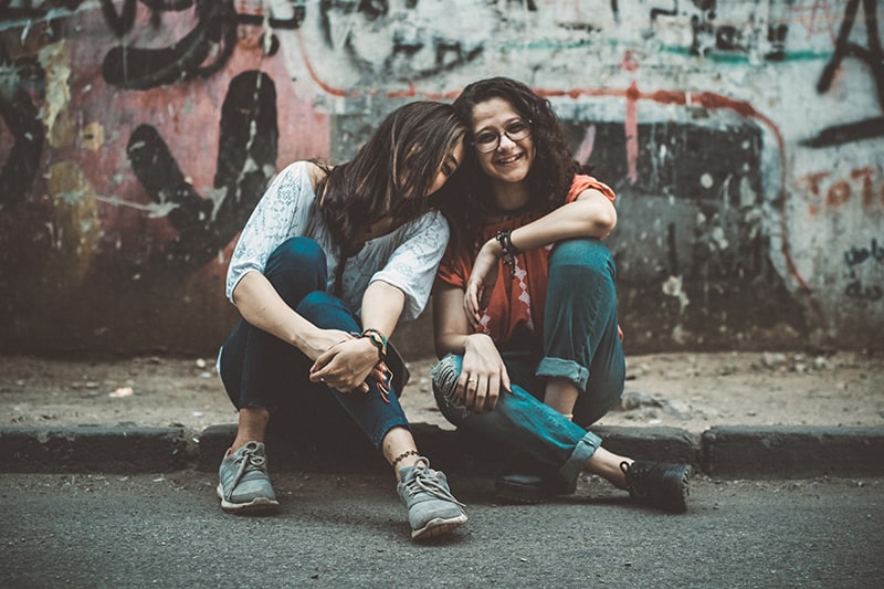 two women sitting on pavement near painted wall