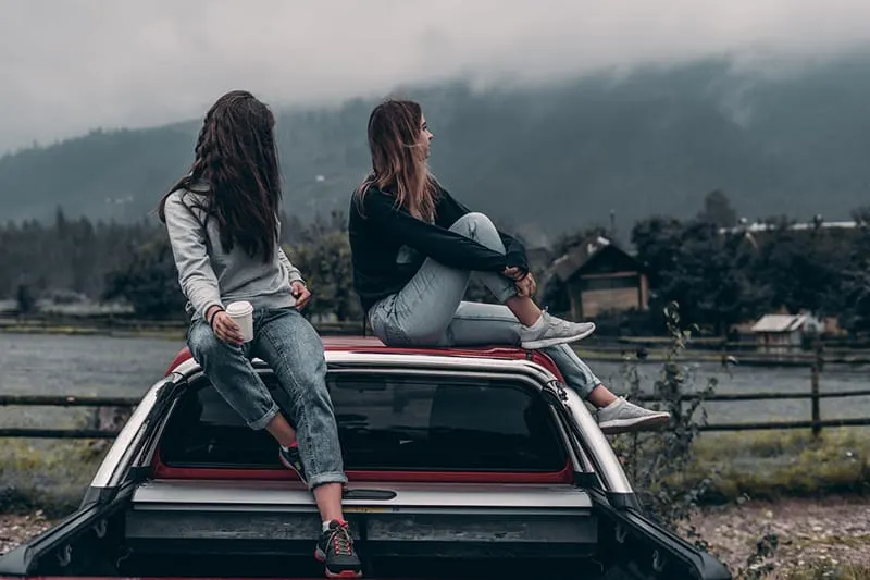two women sitting on vehicle roof near lake