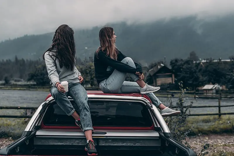 two women sitting on vehicle`s roof near lake