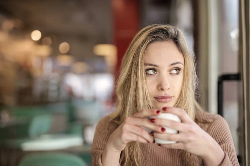 donna che beve da una tazza indossando un lungo top marrone seduta all'interno di un caffè
