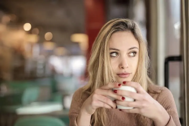 woman drinking from mug wearing long brown top sitting inside a cafe