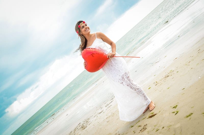 donna che tiene in mano un palloncino a forma di cuore in riva al mare con corona di fiori e abito bianco