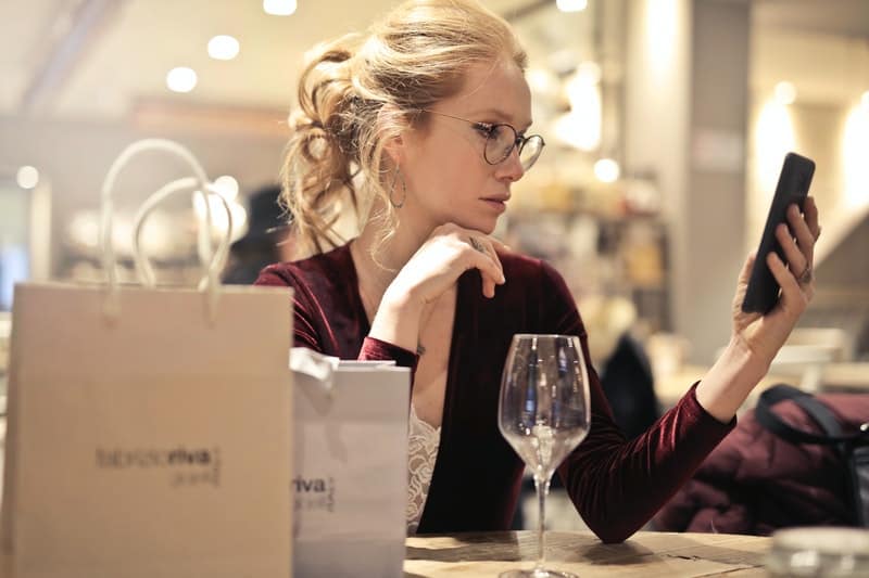 mujer sosteniendo su teléfono con copa de vino sobre la mesa y bolsas de papel