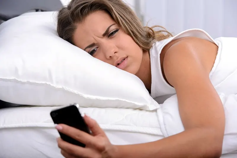 Woman in bed white sheets holding phone looking annoyed 
