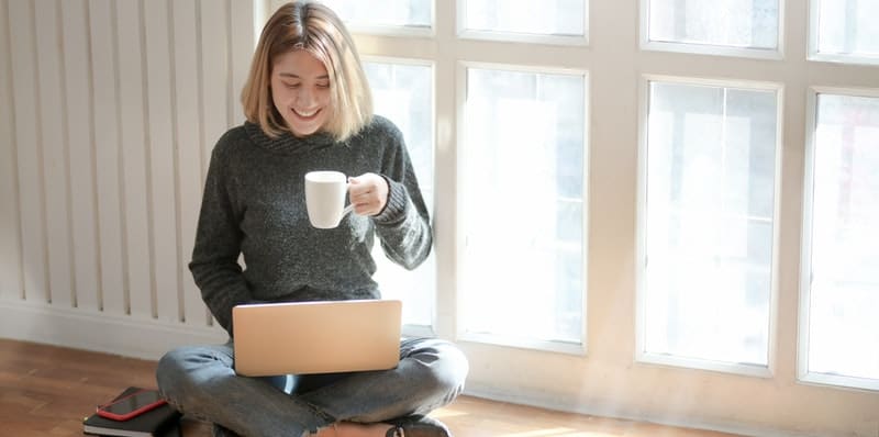 woman in gray sweater drinking coffee sitting on the floor with a laptop