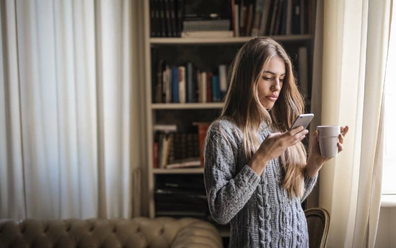 mujer en suéter gris sosteniendo taza de cerámica blanca, mientras que los mensajes de texto en su teléfono