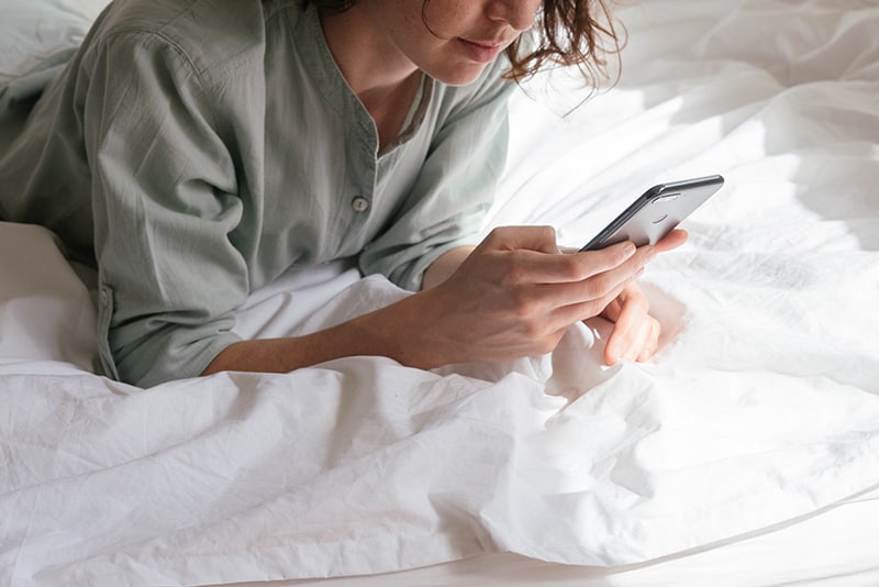 donna in camicia grigia che usa lo smartphone a letto