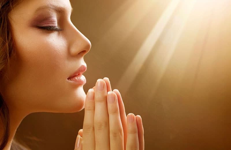 woman in praying hands and eyes closed facing a ray of light