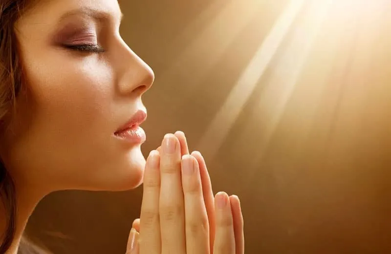 woman in praying hands and eyes closed facing a ray of light
