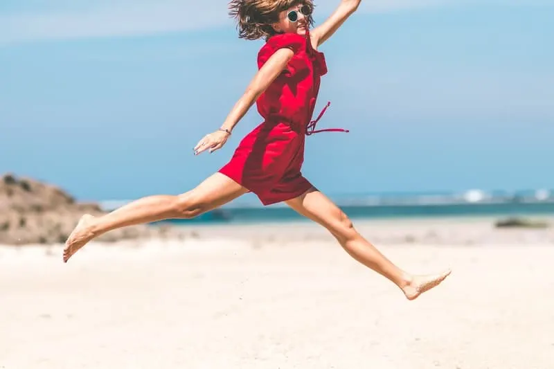 woman in red jumping in the seashore wearing sunglass