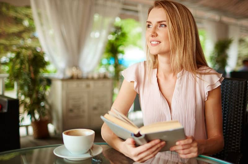 donna in solitudine che sorride tenendo un libro e una tazza sul tavolo 