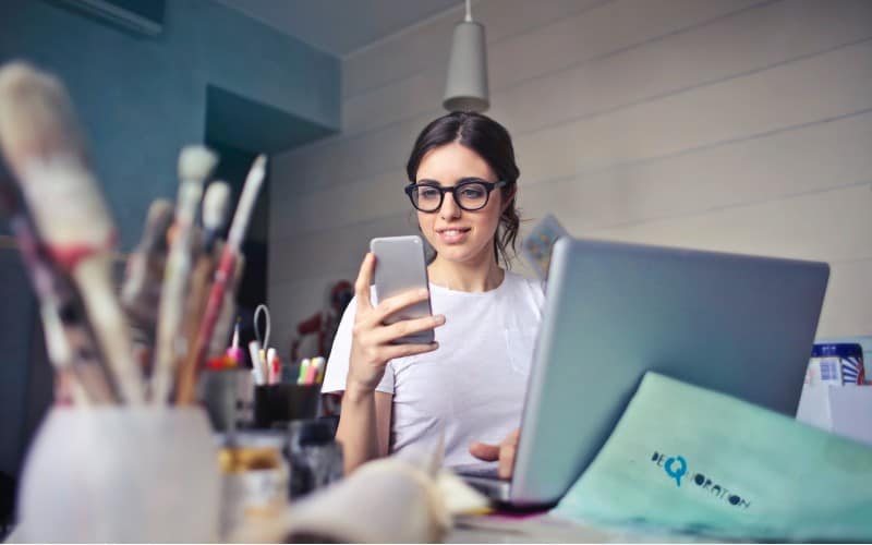woman in white shirt using smartphone while sitting at a desk with laptop on it