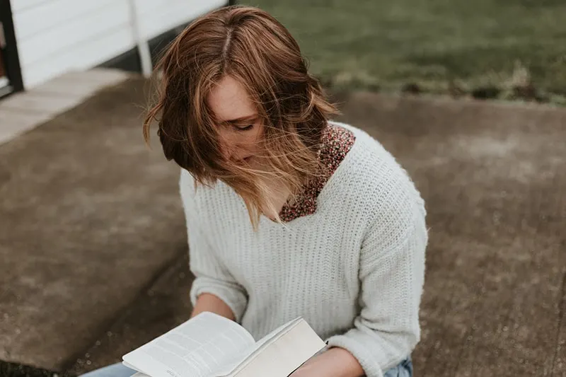 woman in white sweater holding book while sitting on concrete pavement