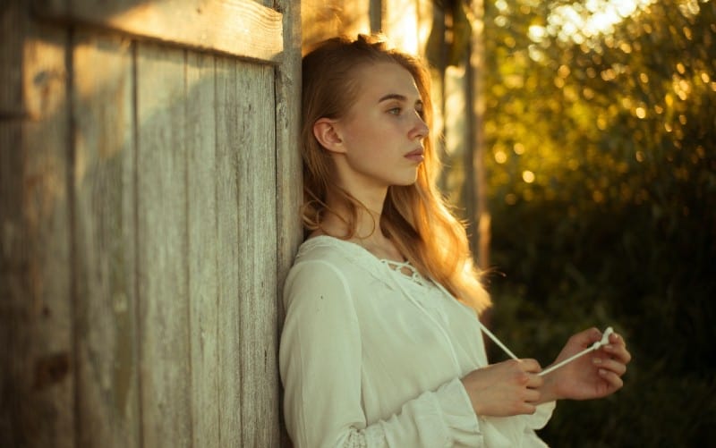 Sad woman wearing white top leaning on wooden wall outdoors