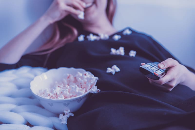 woman lying on bed holding remote control while eating popcorn