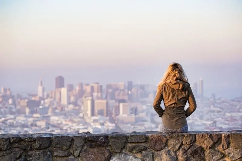 blonde woman sitting on rock looking at city