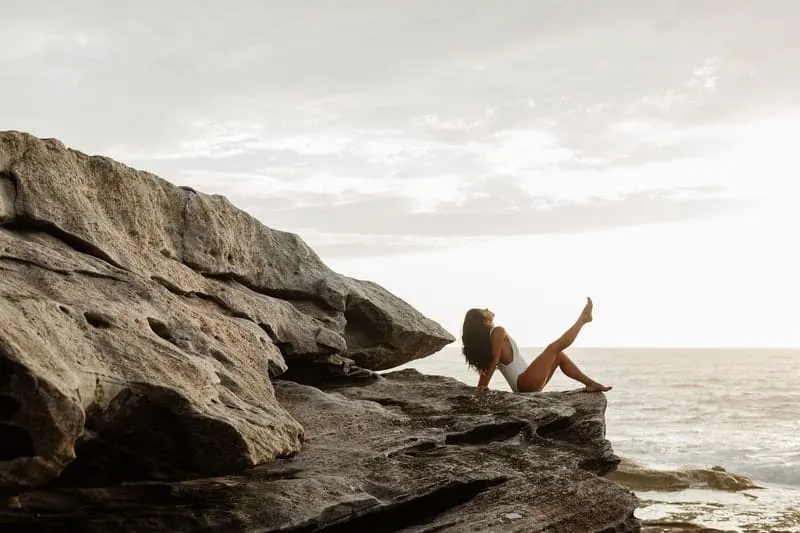 woman on swimwear sitting on rocks near the a body of water with one leg raised