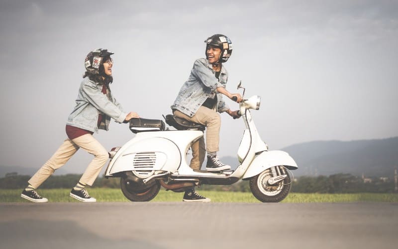 Woman pushing a scooter with man on it druing daytime